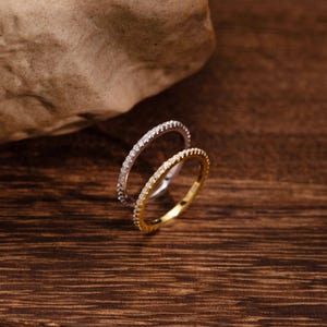 May include: Two eternity rings, one silver and one gold, each set with small, sparkling gemstones. The rings are displayed on a dark wooden surface, with a neutral-toned rock in the background. A classic jewellery design.