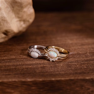 Op de afbeelding: Twee ringen met ovale opaalstenen. Eén ring is zilverkleurig met een halo van kleine, heldere stenen rond de opaal. De andere ring is goudkleurig met een vergelijkbaar halo-ontwerp. Beide liggen op donker hout.