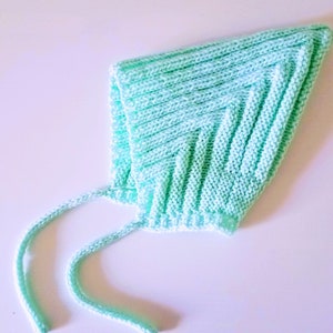 Puede incluir: Un gorro de bebé de punto de color verde claro con una cuerda para atar. El gorro está hecho de un hilo suave y texturizado.