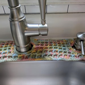May include: A handmade, multi-colored crochet sink skirt, designed to fit around a faucet and soap dispenser. The skirt features a waffle stitch pattern in pastel shades of pink, yellow, blue, and green, adding a decorative touch to the kitchen sink area.