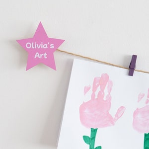 Puede incluir: Una estrella rosa con el texto "Olivia's Art" colgada de una cuerda con una pinza de ropa morada. Una tarjeta blanca con dos flores rosas de huellas de manos con tallos verdes está colgada debajo de la estrella.