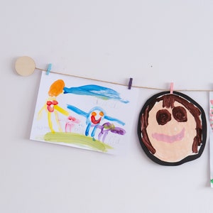 May include: A string of children's artwork hanging on a white wall. The artwork includes a drawing of a sun, a person with outstretched arms, a smiling face, and a tree with pink flowers.