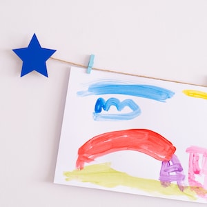 Puede incluir: Una cuerda de coloridas obras de arte de niños está colgada en una pared blanca con acentos de estrellas de papel azul. Las obras de arte representan un arcoíris, nubes, un sol y un árbol.