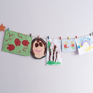 Puede incluir: Cinco dibujos coloridos de niños están colgados en una pared blanca con pinzas de la ropa y una cuerda. Los dibujos incluyen un papel verde con huellas de manos rojas, una cara con cabello castaño, un árbol con flores rosas, dos huellas de manos rojas y un arcoíris.