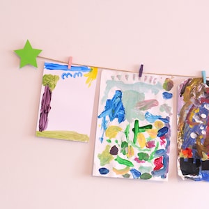 Puede incluir: Tres pinturas coloridas, creadas por un niño, están colgadas en una pared con una estrella verde en la parte superior. Las pinturas están sujetas a una cuerda con pinzas para la ropa.