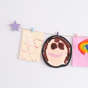 May include: Three children's drawings are hung on a string with clothespins. The drawings are a yellow paper with pink hearts, a black paper with a brown-haired person's face, and a pink paper with a rainbow.