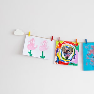 May include: Four pieces of children's artwork are displayed on a white wall using a string and clothes pegs. The artwork includes a pink handprint flower, a colourful abstract painting with a photo of two children in a heart shape, and a blue paper with pink glitter.