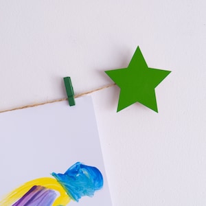 Puede incluir: Un recorte de papel en forma de estrella verde cuelga de una cuerda de yute con una pinza de ropa verde. Una hoja de papel blanca con pinceladas de pintura de colores cuelga debajo de la estrella.