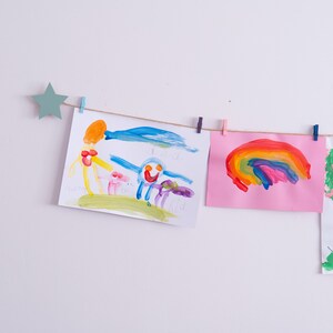 May include: A string of colorful children's artwork is hung on a white wall with clothespins. The artwork includes a rainbow, a tree, and a drawing of a person with a sun and clouds.