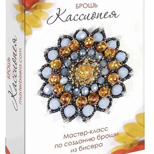 May include: A rectangular box featuring a beaded brooch with a floral design in shades of light blue and amber. The text on the box reads "БРОШЬ Кассиопея" and "Мастер-класс по созданию броши из бисера". The box has a white background with orange scalloped edges.