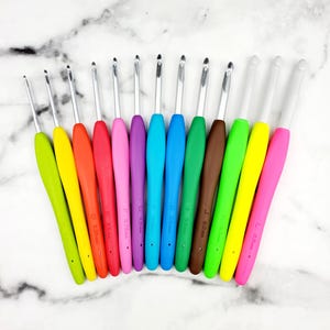 May include: A collection of crochet hooks with colorful, ergonomic handles. The hooks are arranged in a row, showcasing a variety of sizes and vibrant colors, including yellow, red, purple, and blue. Each hook has a silver metal tip.
