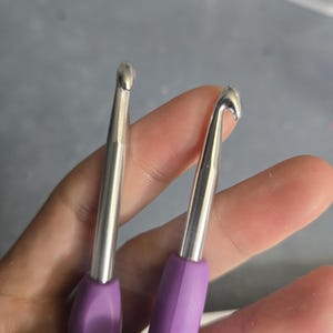 Op de afbeelding: Twee metalen haaknaalden met gladde, zilverkleurige schachten en ergonomische paarse handvatten. De haaknaalden hebben een afgeronde punt en een diepe keel, wat de vorm van de haakkop en de overgang naar het handvat duidelijk maakt.