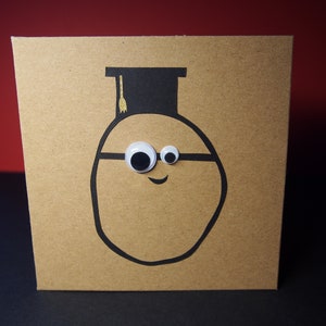 May include: A brown card with a black and white illustration of a potato wearing a graduation cap and glasses. The potato has googly eyes and a smile.