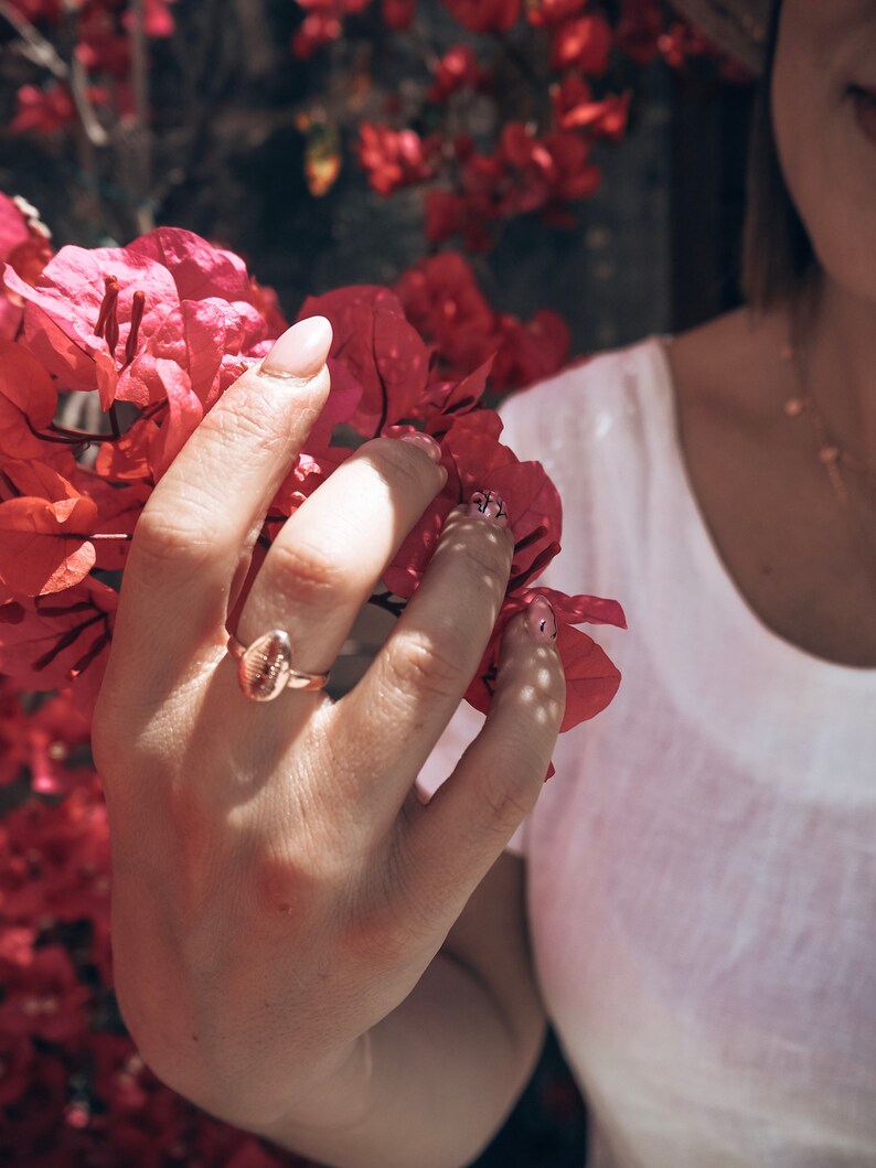 May include: A close-up of a hand wearing a gold ring with a textured oval design. The hand is holding a cluster of pink bougainvillea flowers.