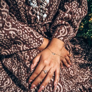 May include: A woman wearing a brown and white patterned dress with a shell necklace and gold rings on her fingers.