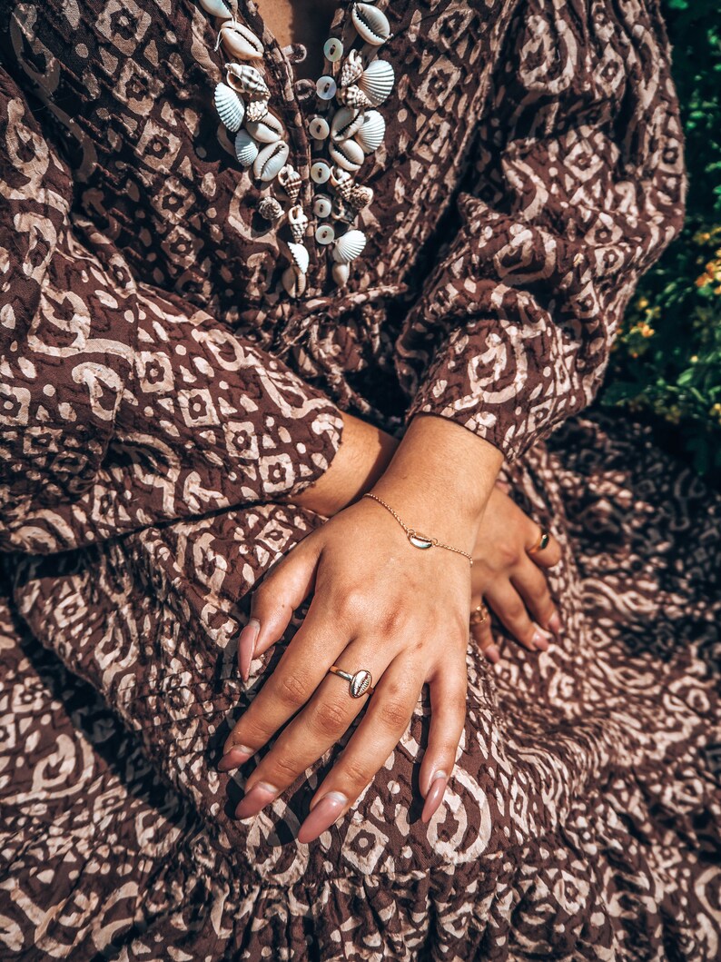 May include: A woman wearing a brown and white patterned dress with a shell necklace and gold rings on her fingers.