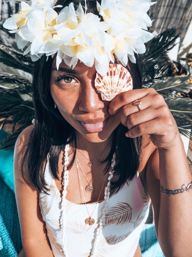 May include: A woman wearing a white flower crown and a white swimsuit with a brown leaf pattern. She is holding a seashell in front of her eye and has a playful expression on her face. She is wearing multiple gold necklaces, including a shell pendant and a coin pendant.