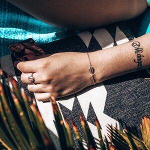 May include: A close-up of a person's arm with a delicate gold bracelet featuring a seashell charm. The bracelet is on a tanned arm with a black tattoo that reads "Mi angel".