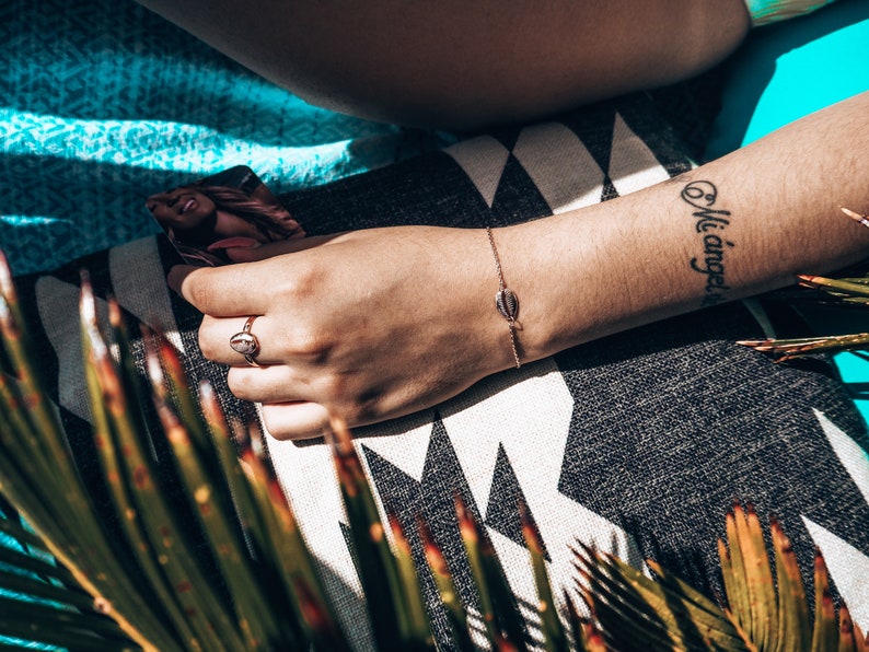 May include: A close-up of a person's arm with a delicate gold bracelet featuring a seashell charm. The bracelet is on a tanned arm with a black tattoo that reads "Mi angel".