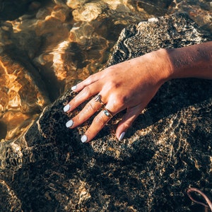 May include: A hand with white nail polish rests on a wet, dark gray rock. The hand is wearing two gold rings.