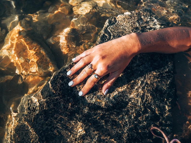 May include: A hand with white nail polish rests on a wet, dark gray rock. The hand is wearing two gold rings.