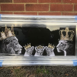 Puede incluir: Una impresión enmarcada en blanco y negro de un león y una leona con coronas doradas y tres cachorros de león con coronas doradas. El fondo es negro.