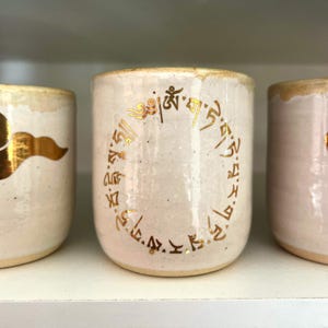 May include: Three handmade ceramic cups with a cream-colored glaze. The central cup features a gold-colored circular inscription. The rims of the cups have a gold-colored trim. The cups are displayed on a white shelf.