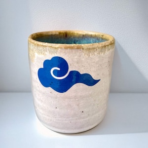 Pode incluir: Um copo de cerâmica com um desenho de nuvem azul. O copo tem um exterior branco-sujo salpicado, uma borda castanha clara e um interior turquesa. A nuvem é uma forma estilizada e rodopiante.