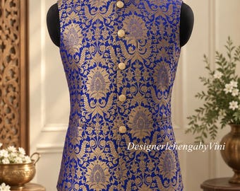 May include: A sleeveless vest in royal blue with a gold floral brocade pattern, featuring a mandarin collar, five gold-toned buttons down the front, and the text DesignerlehengabyVini printed in white on the lower right side of the garment.
