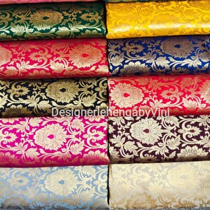 Pu&ograve; includere: Un set di campioni di tessuto di diversi colori e motivi. Il tessuto &egrave; in broccato con un disegno in filo d'oro. I campioni di tessuto sono disposti in fila, con il campione superiore rosa, il secondo verde, il terzo giallo, il quarto blu, il quinto rosso, il sesto nero, il settimo rosa, l'ottavo marrone, il nono azzurro e il decimo beige. Il testo "DesignerlehengabyVini" &egrave; visibile sui campioni di tessuto.