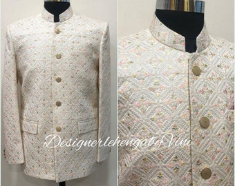 Pode incluir: Casaco cor marfim com padrão geométrico e botões dourados. O casaco tem gola mandarim e dois bolsos frontais. O tecido é bordado com um desenho floral em tons de rosa e verde. O texto "Designerlehengaby Vini" é visível.