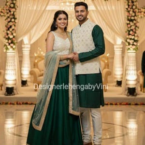 Puede incluir: Una pareja posa con ropa formal. La mujer lleva una falda verde y un top blanco con detalles dorados, complementado con un chal verde transparente. El hombre viste una túnica verde, un chaleco color crema y pantalones blancos. El fondo presenta decoraciones florales y telas drapeadas.