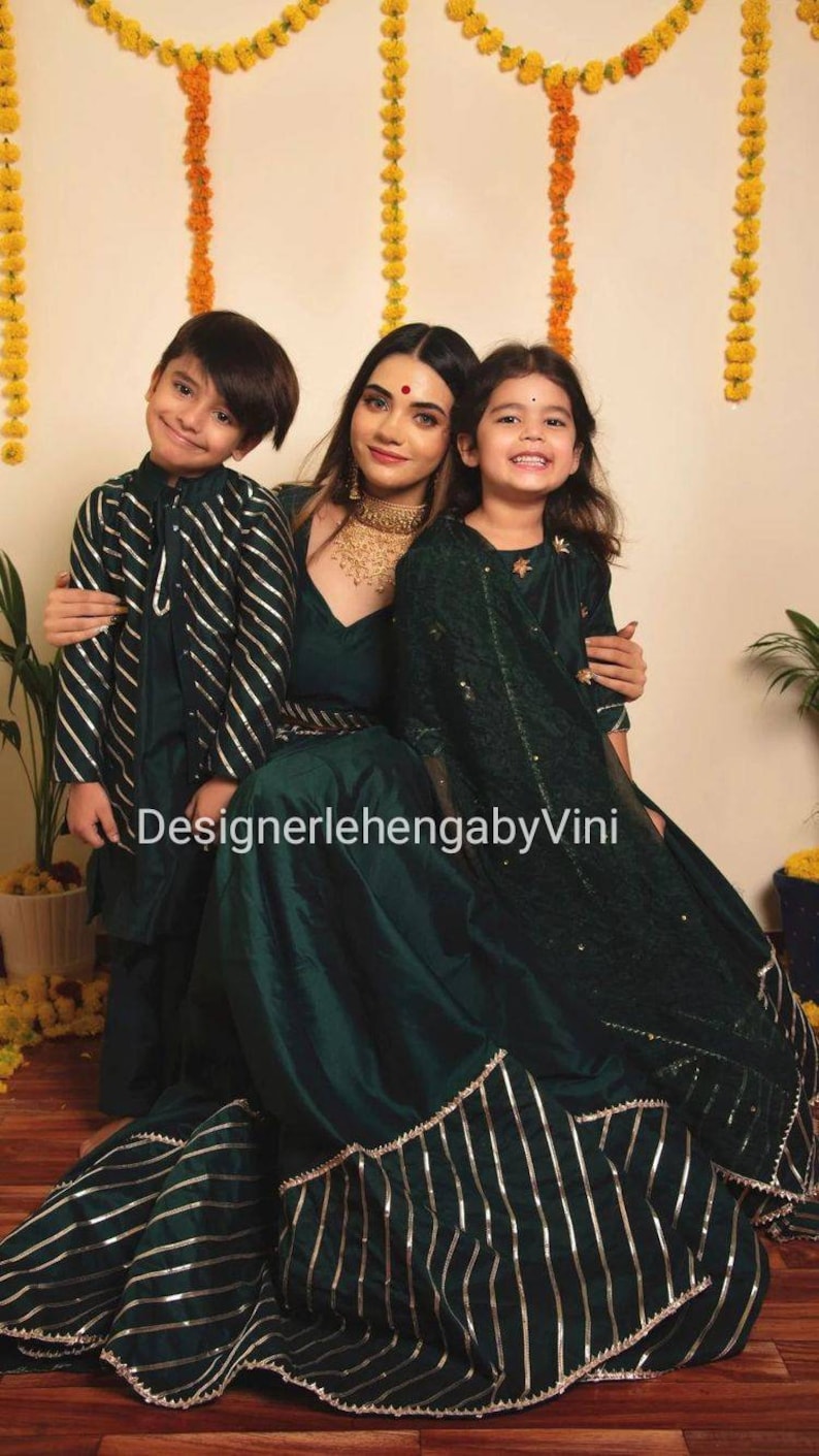 May include: A family of three wearing matching green outfits with silver and gold accents. The woman is wearing a long green dress with a silver and gold striped trim. The two children are wearing matching green shirts with silver and gold stripes. The background is a light yellow wall with yellow flower garlands hanging from the ceiling. The text "DesignerlehenagabyVini" is visible in the image.