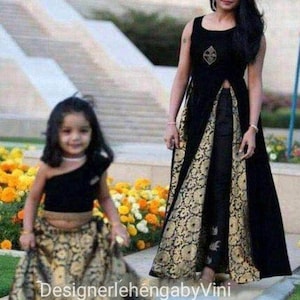 Op de afbeelding: Een moeder en dochter dragen bijpassende zwarte en gouden outfits. De moeder draagt een lange zwarte jurk met een gouden brokaatrok en een zwart topje. De dochter draagt een zwart crop topje en een gouden brokaatrok. De tekst "DesignerlehengabbyVini" is zichtbaar onderaan de afbeelding.