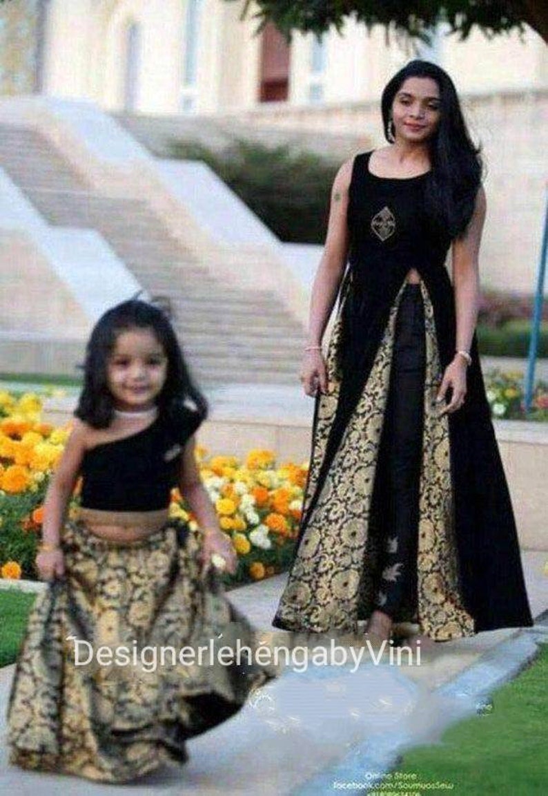 Op de afbeelding: Een moeder en dochter dragen bijpassende zwarte en gouden outfits. De moeder draagt een lange zwarte jurk met een gouden brokaatrok en een zwart topje. De dochter draagt een zwart crop topje en een gouden brokaatrok. De tekst "DesignerlehengabbyVini" is zichtbaar onderaan de afbeelding.