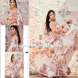 May include: A woman wearing a white and pink floral print lehenga with a matching crop top. The lehenga has a flowing skirt and the crop top has a sleeveless design. The woman is wearing her hair down and has a gold necklace on. The image also includes the text "Designerlehenga by Vini".