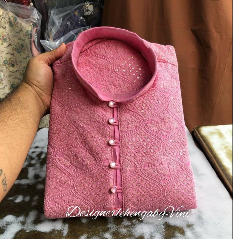 Puede incluir: Una kurta rosa con cuello alto y bordados intrincados. La kurta tiene cierre de botones y est&aacute; decorada con peque&ntilde;as cuentas blancas. El texto "DesignerlehengabyVini" es visible en la parte inferior de la imagen.