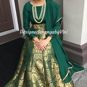 Könnte beinhalten: Ein grünes und goldenes Brokat-Lehenga mit passendem Dupatta. Das Lehenga hat ein tailliertes Oberteil und einen fließenden Rock. Das Dupatta ist mit einem goldenen Rand verziert. Der Text "DesignerlehengabyVini" ist auf dem Bild sichtbar.