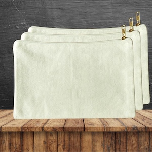 Könnte beinhalten: Vier cremefarbene Canvas-Reißverschlusstaschen mit goldfarbenen Reißverschlüssen. Die Taschen sind rechteckig und scheinen aus einem strukturierten Stoff zu bestehen. Sie sind leicht überlappend auf einer Holzoberfläche angeordnet.