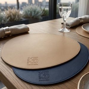 May include: Two round placemats, one tan and one navy blue, on a wooden table. The tan placemat is on top of the navy blue one. Each placemat has the text "BRISA 35 ROOFTOP DINING". A glass of water, a plate, and a napkin are also on the table.