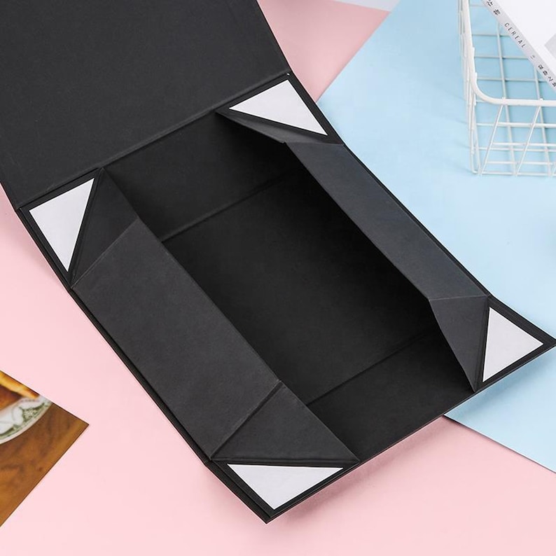 May include: Black folding gift box with white triangular accents on the inside flaps. The box is open, revealing the interior, and is set against a pink and blue background. The box is made of a matte material.