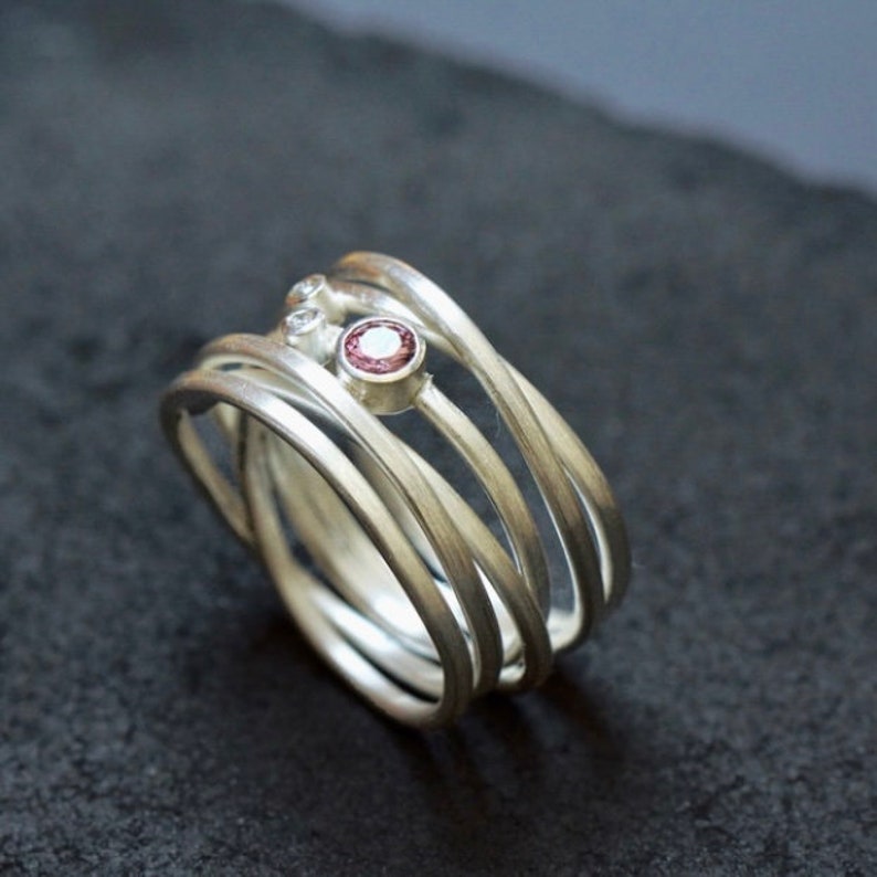 May include: A silver ring with five bands, each with a slightly different width. The bands are twisted and intertwined, and there are three small gemstones set into the ring. The center stone is a pink gemstone, and the two smaller stones are white.