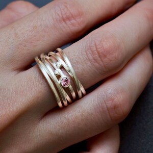 May include: A gold ring with multiple bands and three gemstones. The ring features a pink gemstone and two smaller white gemstones.