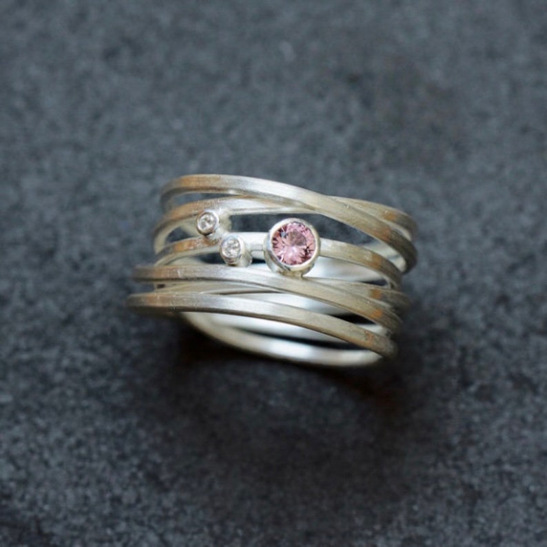 May include: A silver ring with multiple bands, featuring a pink gemstone and two smaller clear gemstones.