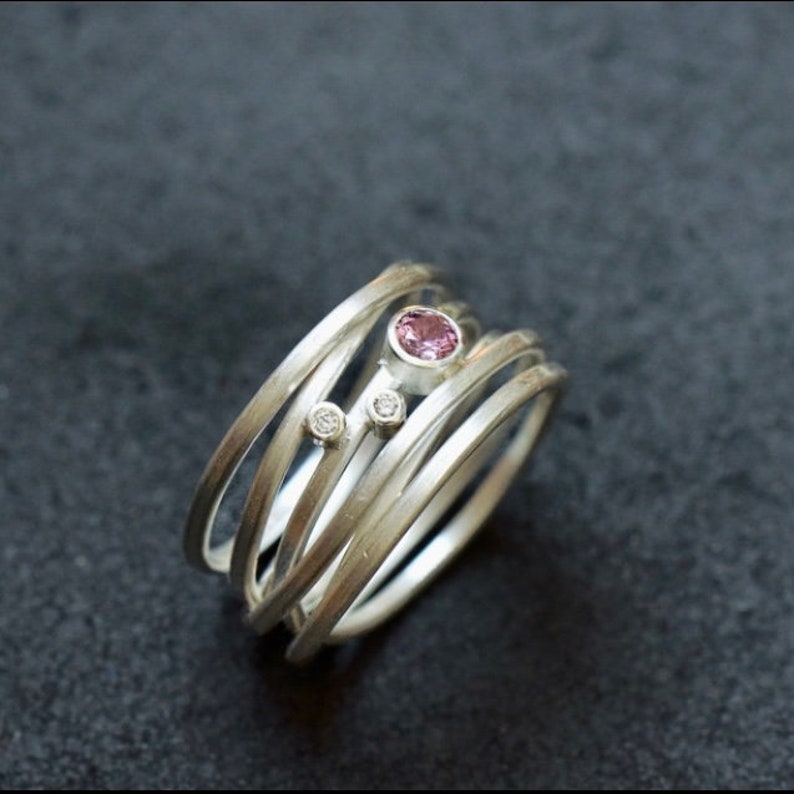 May include: A silver ring with multiple bands, featuring a pink gemstone and two smaller white gemstones.