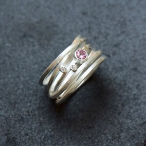 May include: A silver ring with multiple bands, featuring a pink gemstone and two smaller white gemstones.