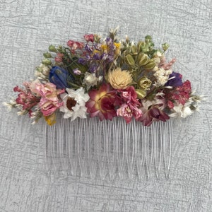 May include: A hair comb decorated with a variety of dried flowers in shades of pink, purple, blue, yellow, and green. The comb is made of clear plastic and has a row of teeth.
