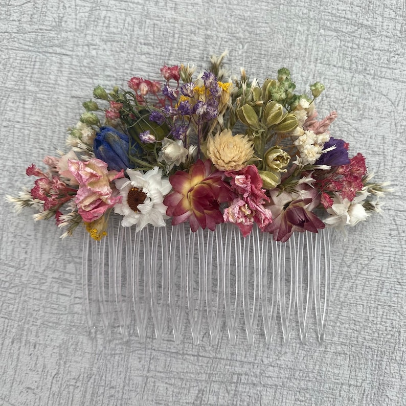 May include: A hair comb decorated with a variety of dried flowers in shades of pink, purple, blue, yellow, and green. The comb is made of clear plastic and has a row of teeth.