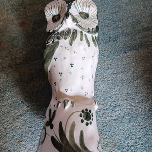 May include: A white ceramic owl ornament with hand-painted details. The owl has large eyes and is decorated with green and black floral and dot patterns. The figurine has a tall, slender shape, likely for decorative purposes.