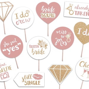May include: A set of 10 pink and gold glitter photo booth props for a bridal shower or bachelorette party. The props feature phrases like "I do crew", "bride squad", "miss to Mrs", "she said yes", "team bride", "but first, #selfie", "still single", "already taken", "I do", and "cheers".